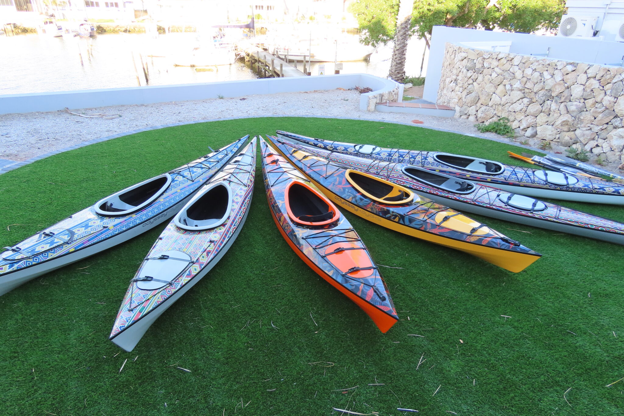 Variety of Odyssey kayaks all aligned on the grass overlooking a marina with yachts